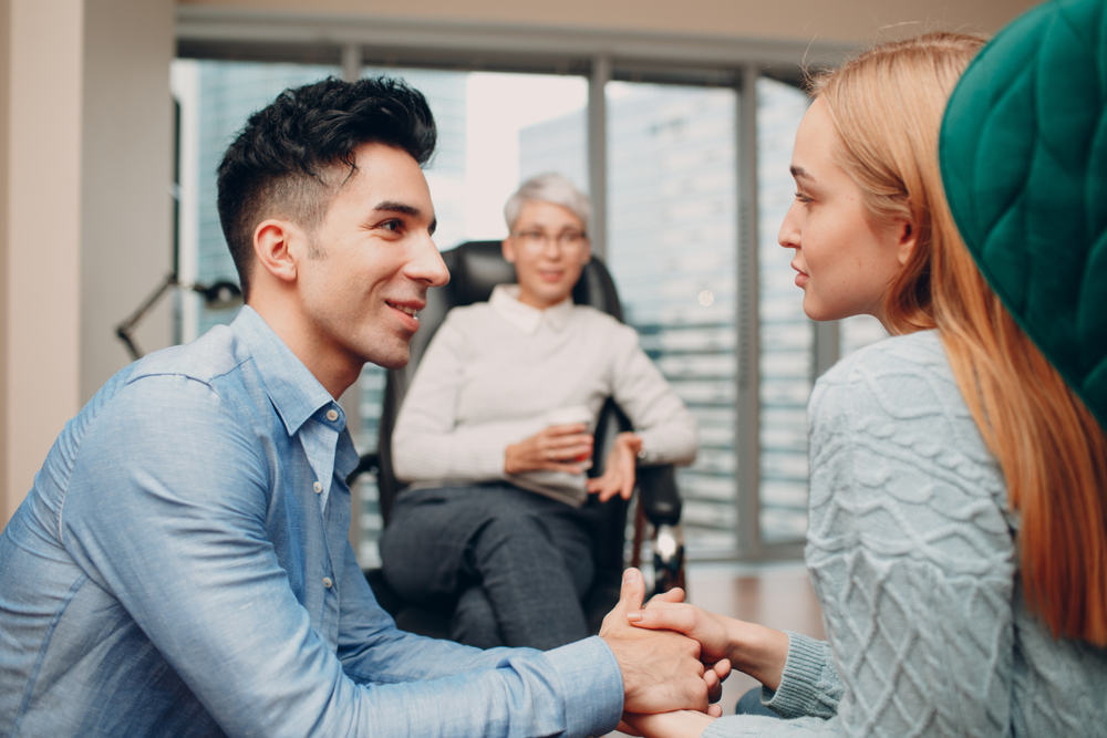 A man and a woman sit facing each other holding hands while a counselor listens from a chair in the background.