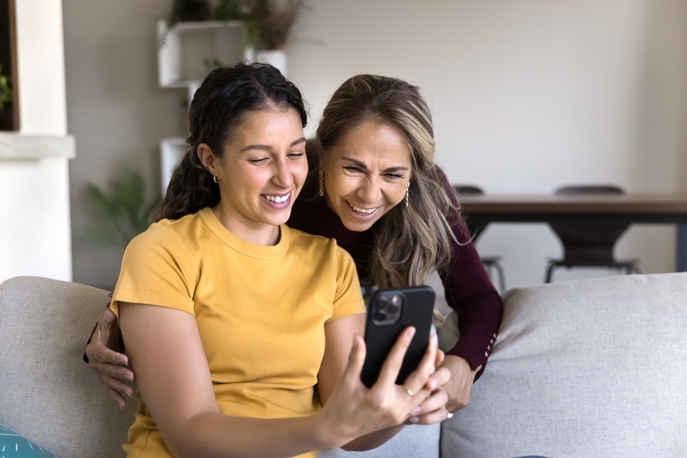 Mother and her adult daughter laughing together while looking at a phone on a couch at home