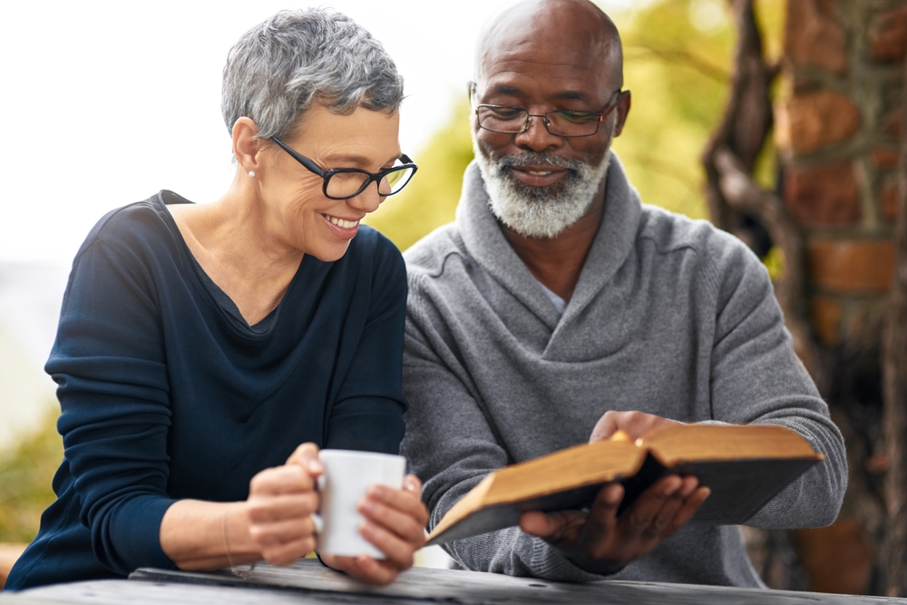 An older man and woman sit together at an outdoor wooden table, smiling, as he holds an open book and she holds a white mug.