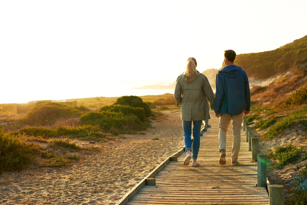 A man and a woman hold hands as they walk away from the camera along a wooden boardwalk through coastal dunes at sunset.